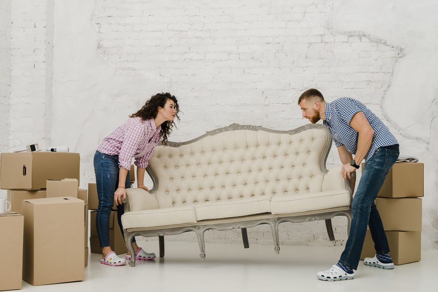 Two people lifting a large sofa in a room stacked with moving boxes